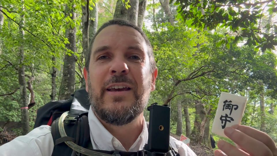 The hiker, smiling into his phone camera, holds up a small wooden omamori charm.