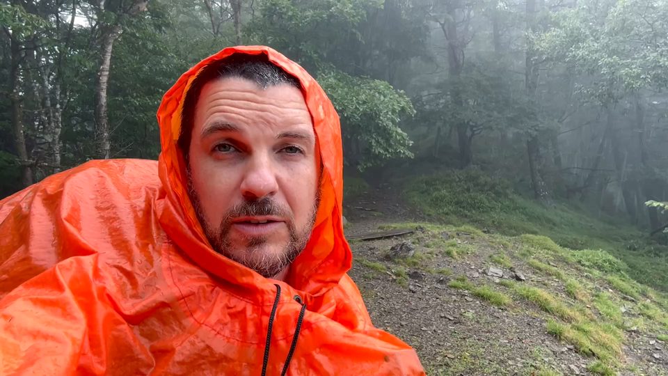 The hiker in a blaze-orange rain poncho, hood up, in a foggy forest.