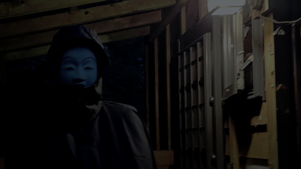 Nightfall under a wooden hut's covered walkway: a hooded figure in a pale Noh mask stands beside a glowing lantern.