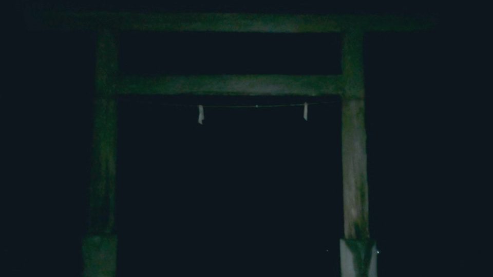 A wooden torii gate at night, barely lit; between its posts, a shimenawa rope with paper streamers hangs in the dark.